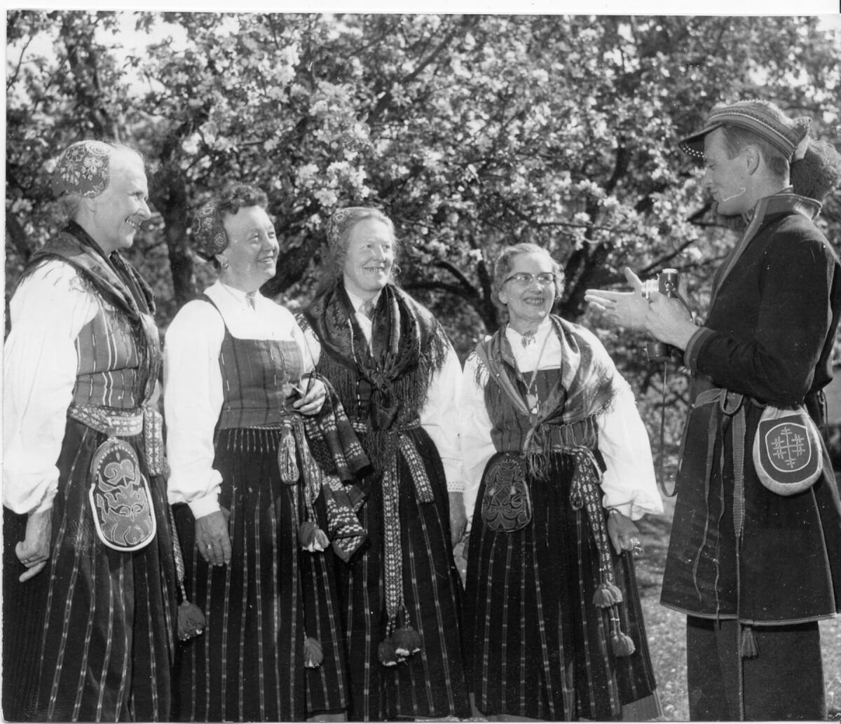 Fyra kvinnor står framför en man med kamera. Kvinnorna har folkdräkt, Delsbodräkt med sjalar, hättor och väskor. Mannen har klätt sig som en same och håller i en småbildskamera. Det är sommar och de står framför ett blommande äppelträd.