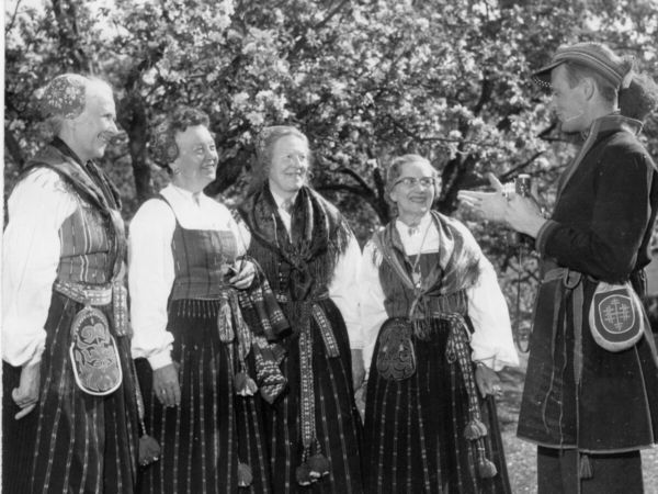 Fyra kvinnor står framför en man med kamera. Kvinnorna har folkdräkt, Delsbodräkt med sjalar, hättor och väskor. Mannen har klätt sig som en same och håller i en småbildskamera. Det är sommar och de står framför ett blommande äppelträd.