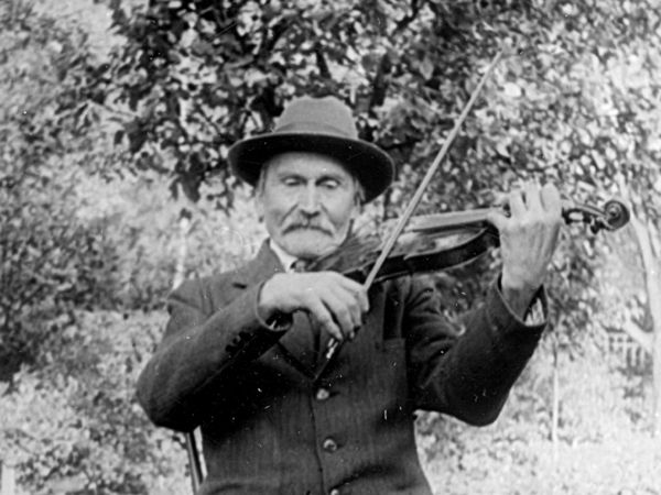 Bild på spelmannen Karl Herman Erlandsson som sitter och spelar fiol på en stol, utomhus.