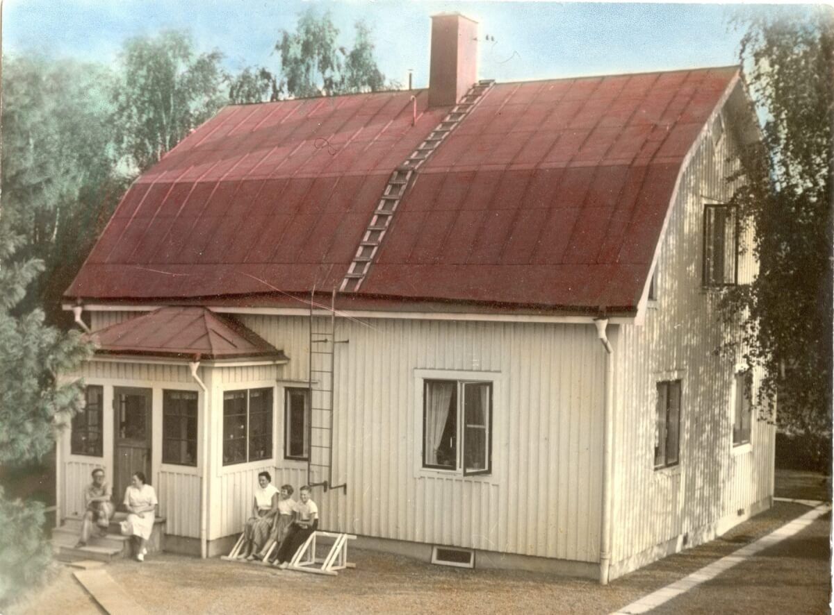 Ett hus med plåttak och träfasad. Framför huset sitter fem personer.