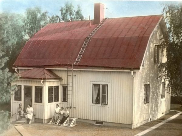 Ett hus med plåttak och träfasad. Framför huset sitter fem personer.