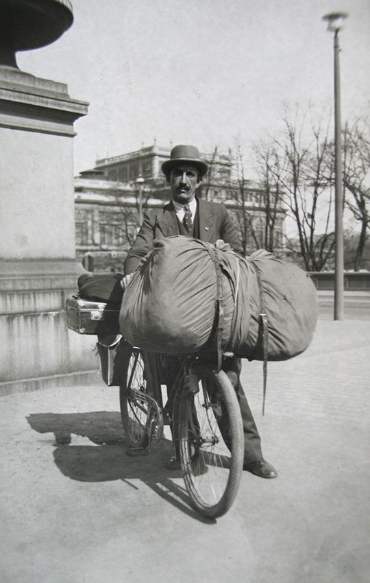Svartvitt fotografi på en man som står med sin välpackade cykel. Mannen är klädd i kostym och bär hatt.