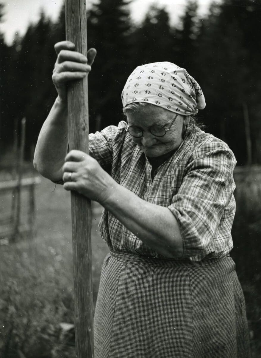 En gammal kvinna håller upp en kraftig trästång med bägge händerna. Hon har runda glasögon, ett huckle över håret, smårutigt förkläde. Man ser att hon håller stången i ett fast grepp med sina starka händer. Bilden är tagen på landet, man ser en mörk skog i bakgrunden.