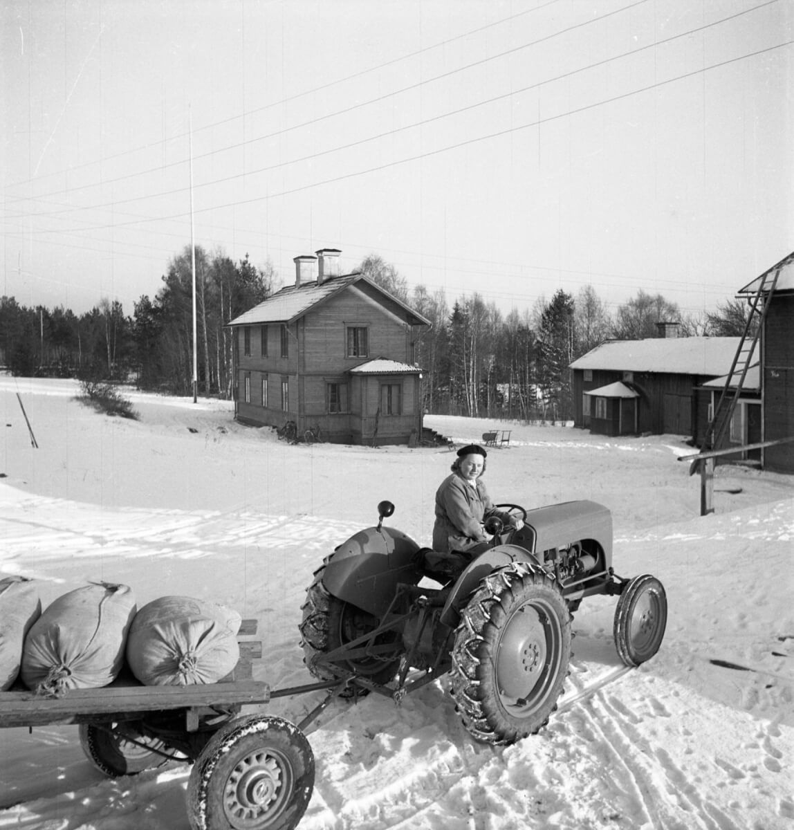 En kvinna kör en traktor som drar ett släp med några säckar.