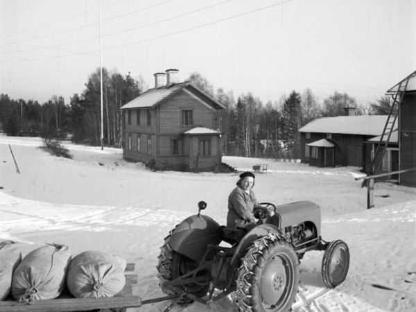En kvinna kör en traktor som drar ett släp med några säckar.