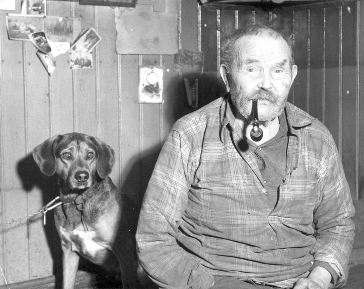 Porträtt av en man som röker pipa och en hund sitter bakom honom.