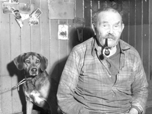 Porträtt av en man som röker pipa och en hund sitter bakom honom.