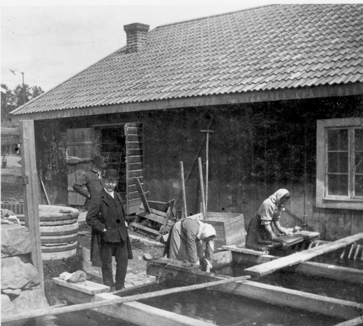 Två kvinnor arbetar intill en fiskebod i skärgården. Två män i kostym och hatt ser på.