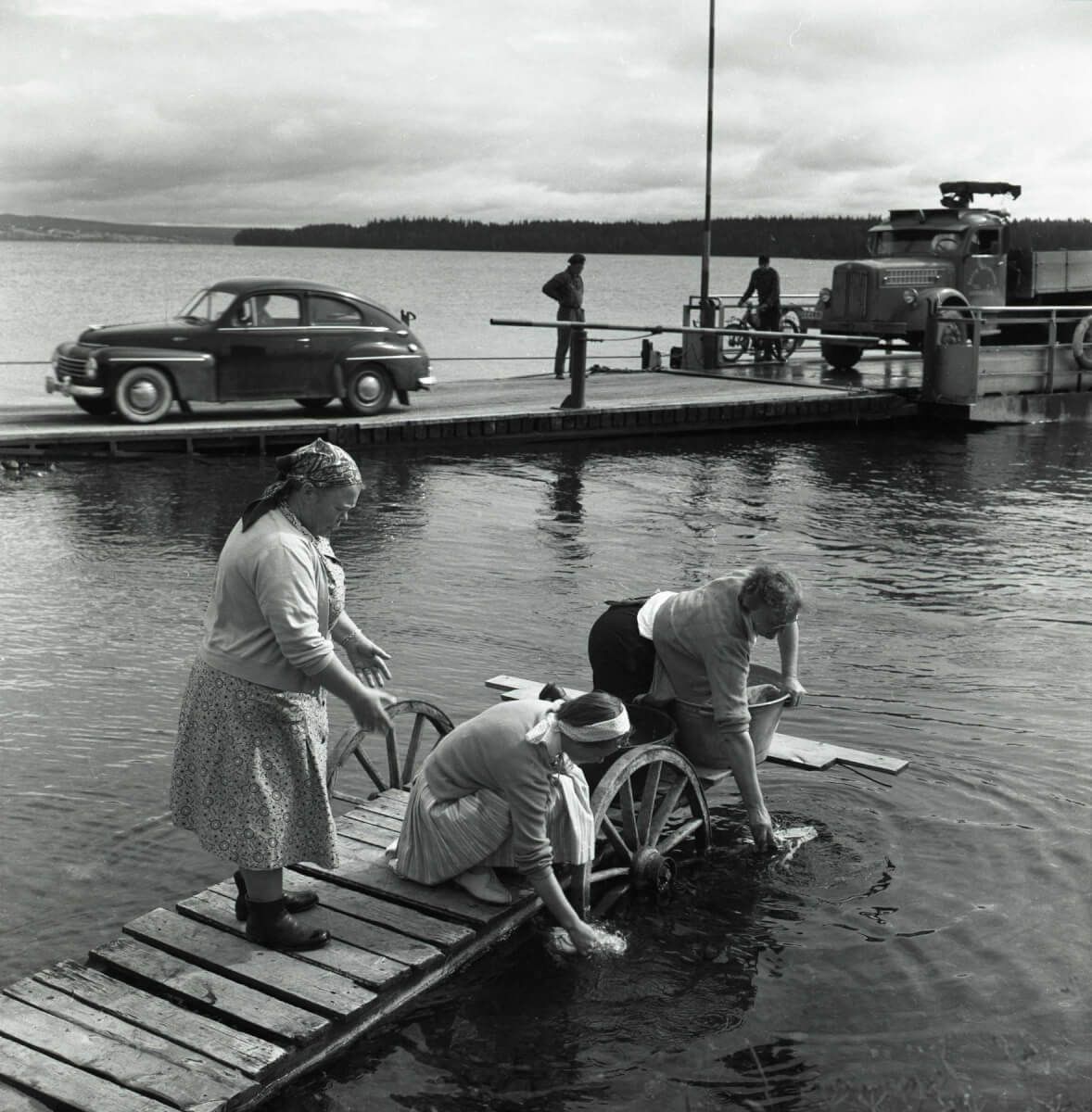 Tre kvinnor tvättar kläder från en brygga. I bakgrunden har en bilfärja dockat och fordonen kör av.