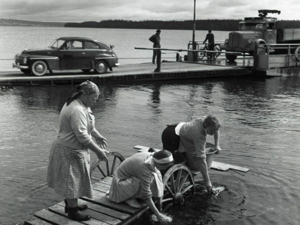 Tre kvinnor tvättar kläder från en brygga. I bakgrunden har en bilfärja dockat och fordonen kör av.