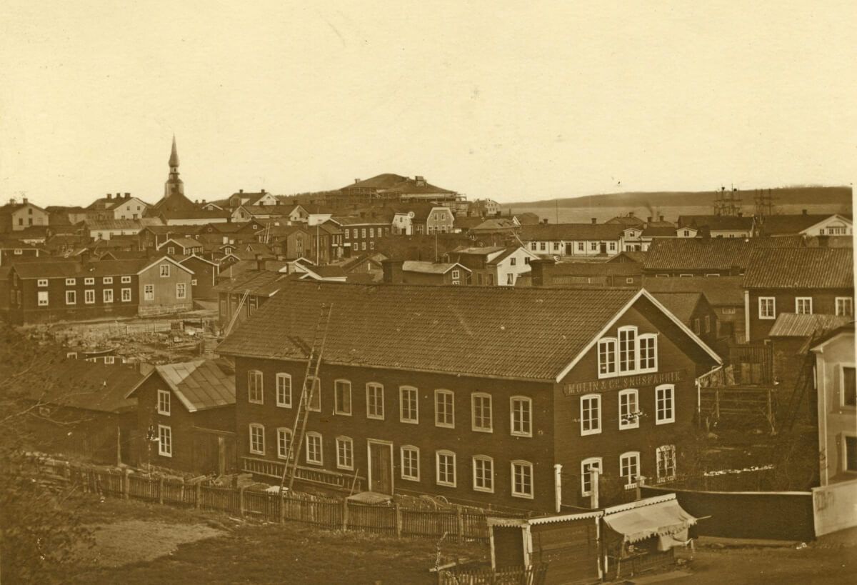 En mycket gammal bild av Hudiksvall med stora och små trähus som trängs längs gatorna. En stor träbyggnad i förgrunden har en skylt på gaveln där det står Molin & Co Snusfabrik. Man kan se Hudiksvallsfjärden och ett segelfartyg. Det gamla rådhuset sticker upp sin takspira till vänster. Bilden är tagen för 1879.
