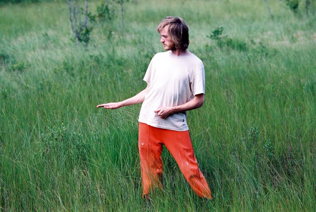 En man iförd orangea byxor och vit t-shirt står i gräset.