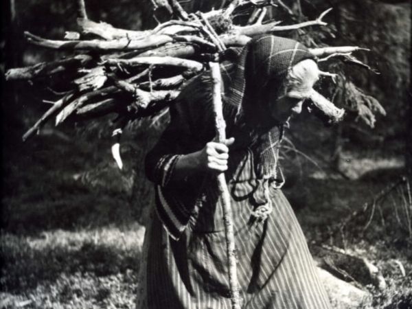 En äldre kvinna bär ved på ryggen och stödjer sig på en pinne med ena handen.